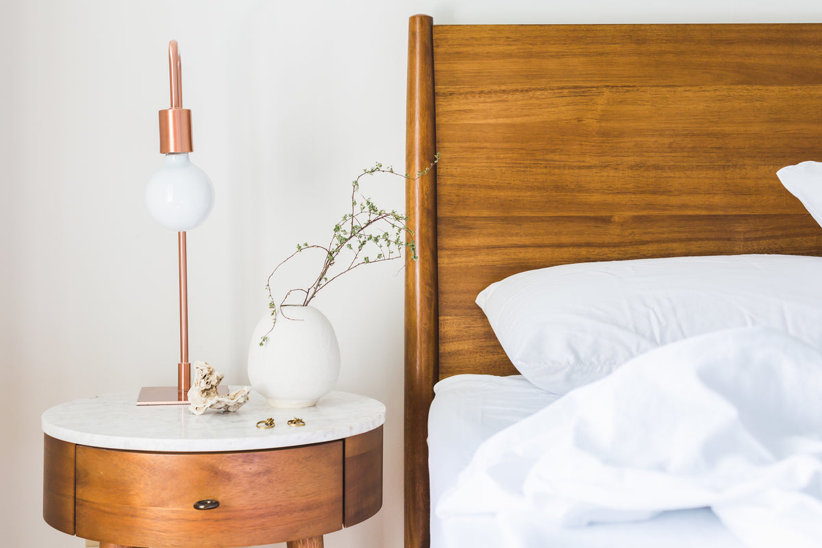 Warm, sunlit bedroom corner with a wood bed frame, copper lamp and cozy linens, The Goods Nook homepage hero
